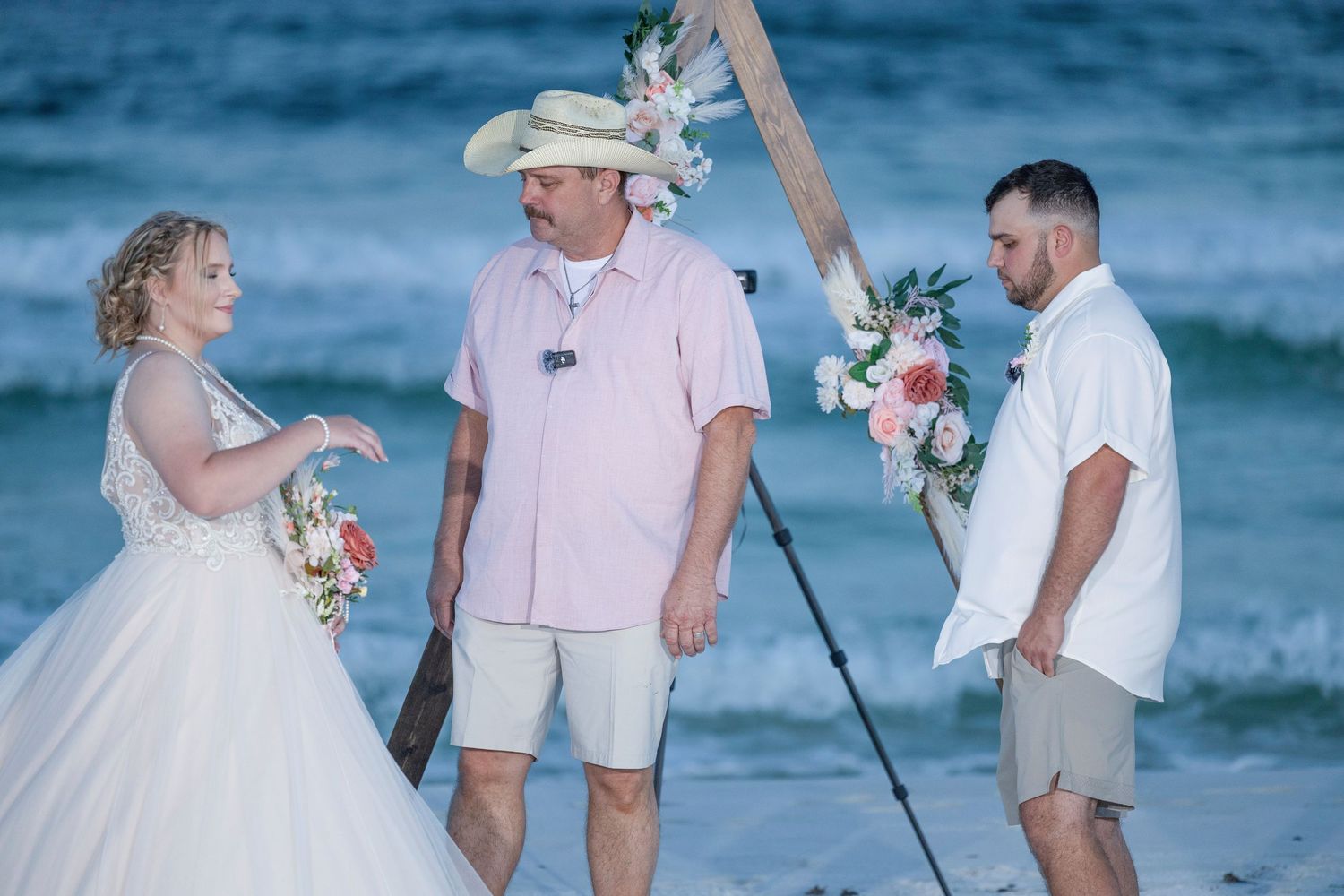 A beach wedding ceremony takes place at dusk with people gathered by the ocean under soft blue lighting.