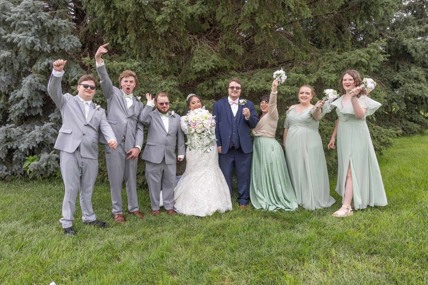 Wedding party members pose playfully on a grassy lawn with trees in the background during outdoor celebration.