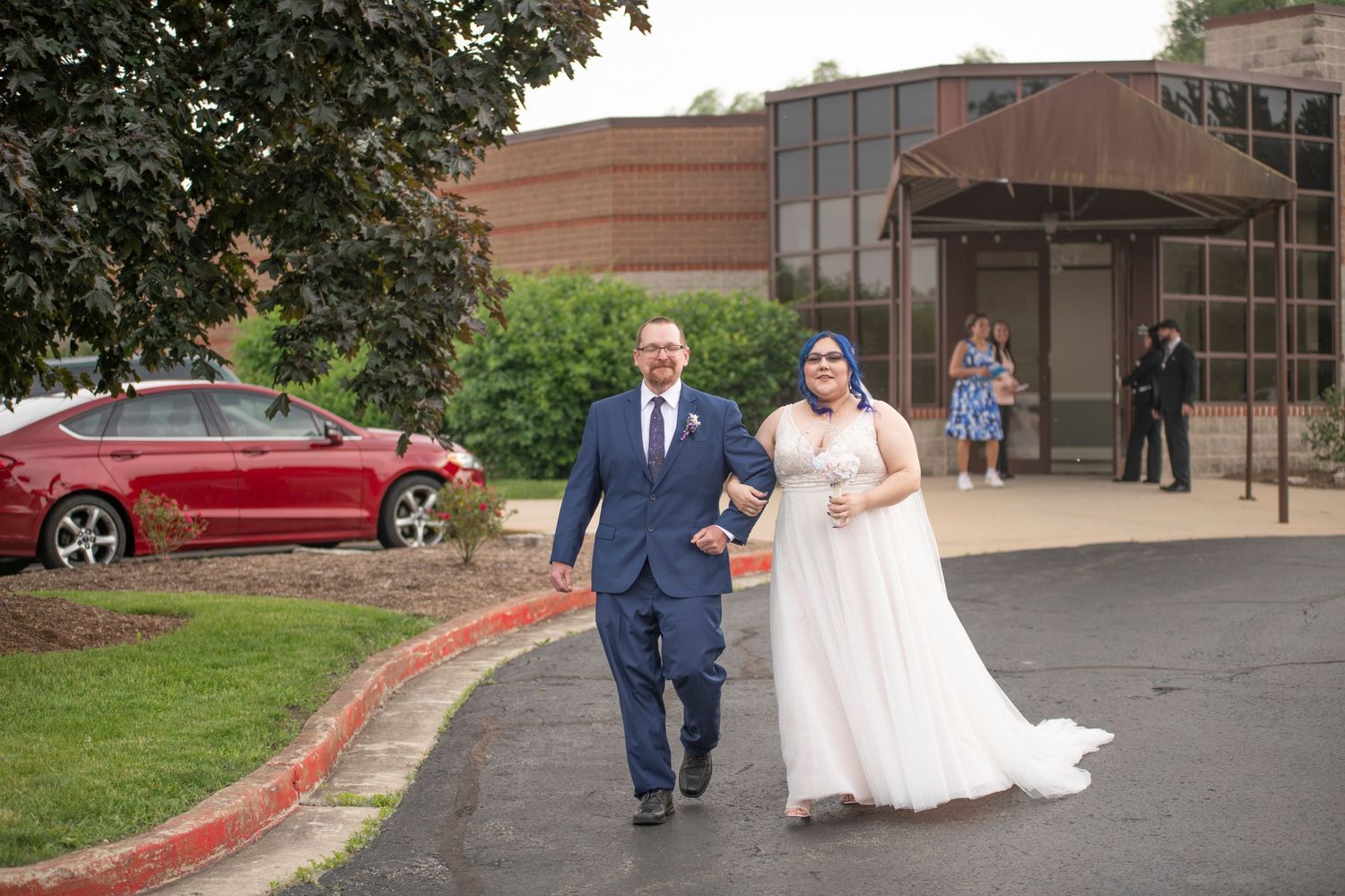 A wedding couple walks together in a parking lot surrounded by brick buildings and greenery.