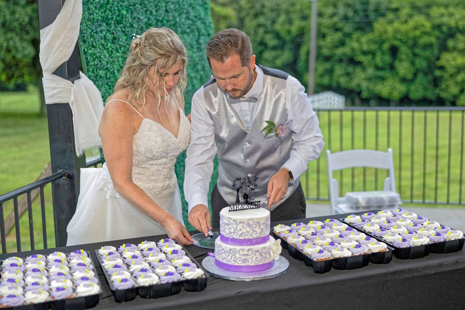 A couple cutting their wedding cake together at an outdoor reception with cupcakes displayed on black tables.