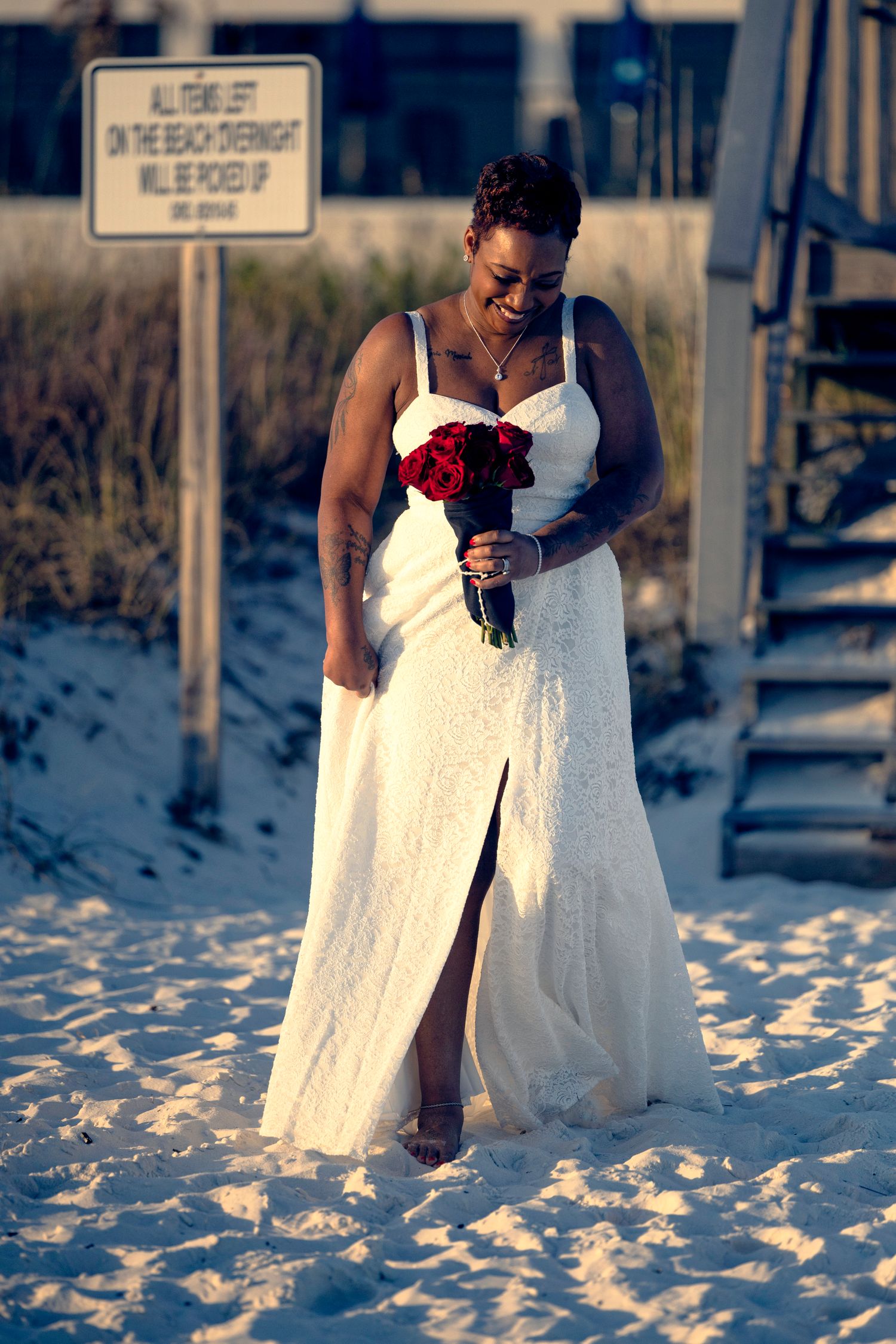 A bride in a white lace wedding dress walks along a snowy beach holding a red rose bouquet.