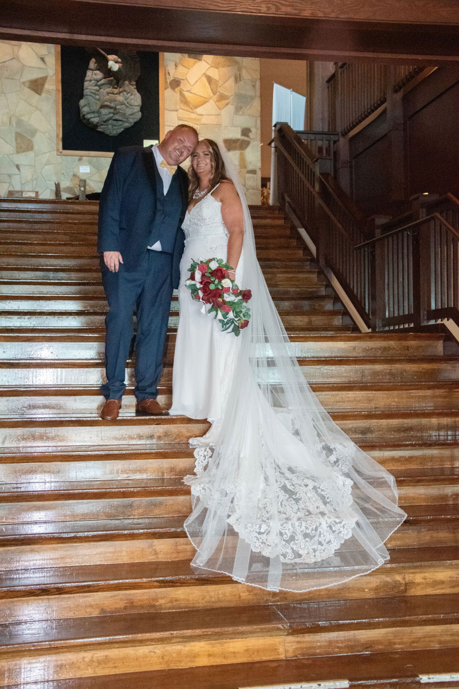A couple stands on wooden stairs during their wedding ceremony, with the bride's lace dress train elegantly cascading down.