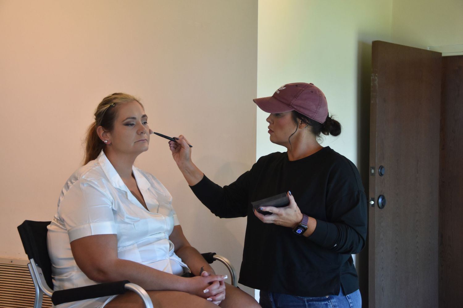 A makeup artist applies cosmetics to a client seated in a chair while holding a makeup palette.
