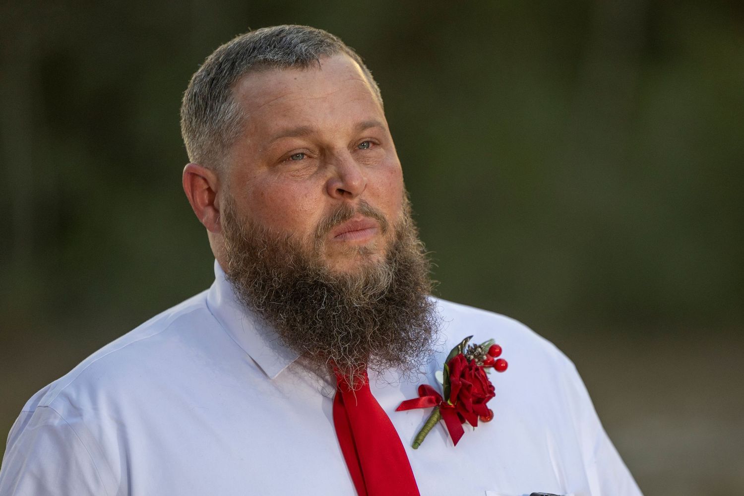 A person in a white shirt and red tie wearing a red carnation boutonniere with a full beard.