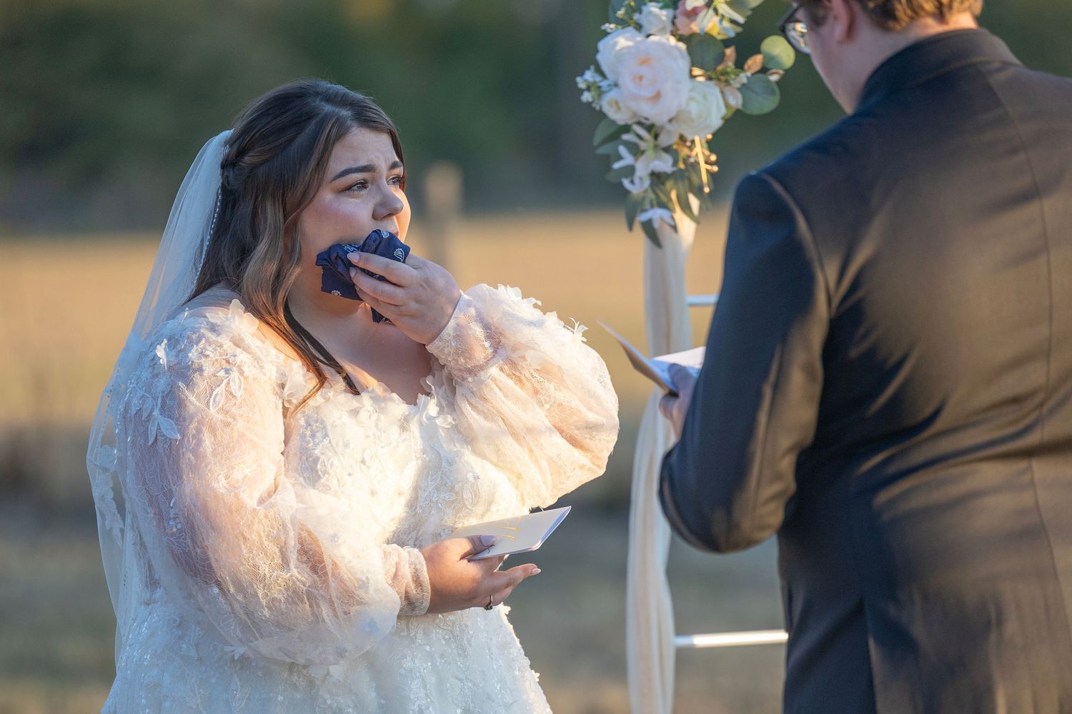 Bride in a white lace gown laughs joyfully during an outdoor wedding ceremony at sunset.