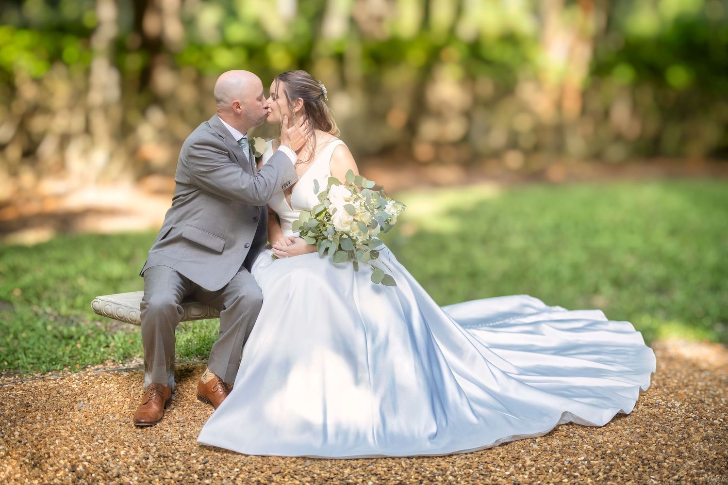 A romantic wedding photo outdoors shows a couple in formal attire sharing a tender moment on a sunlit garden path.