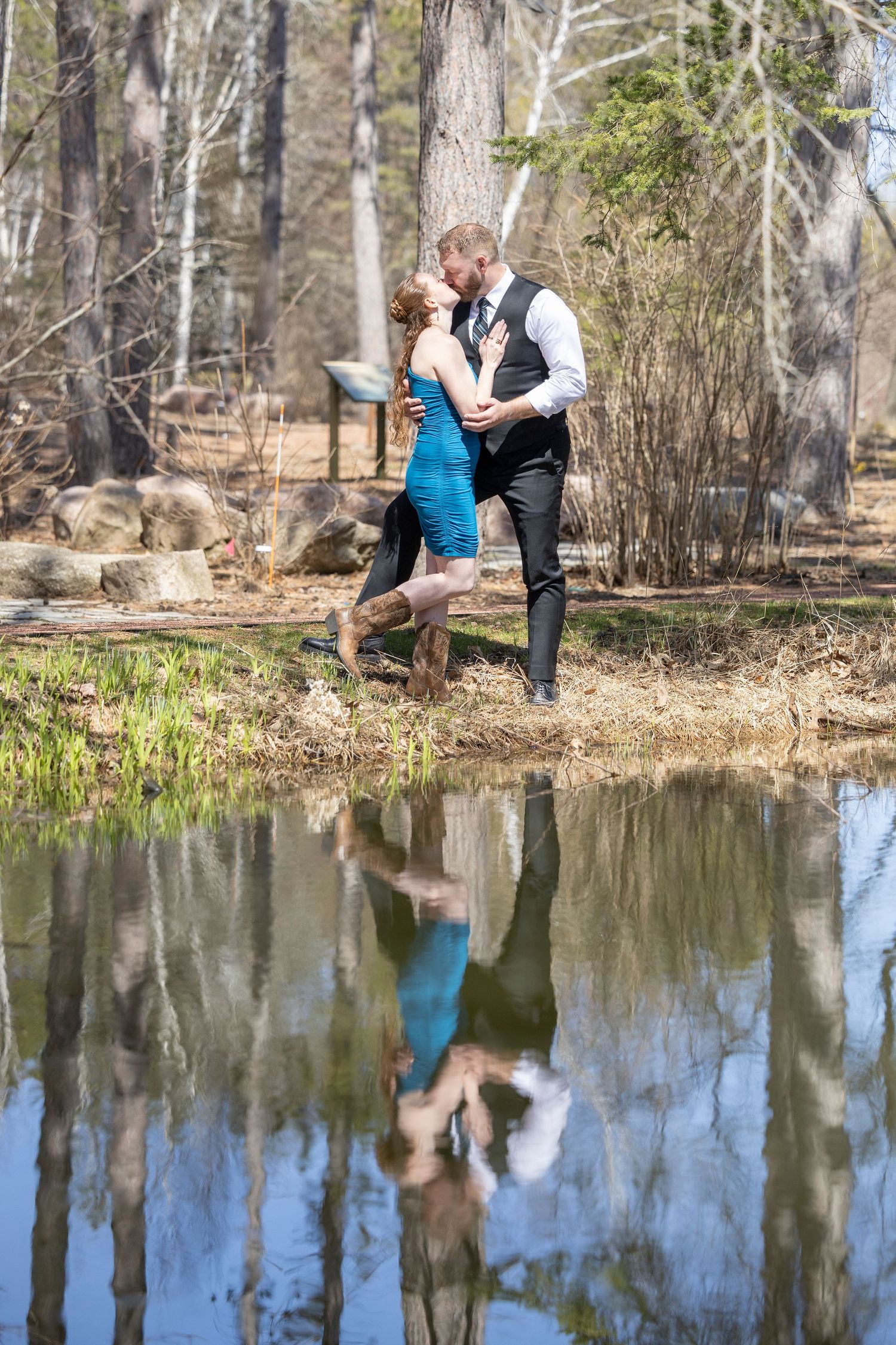 A couple shares a romantic moment by a reflective pond in a wooded park setting.