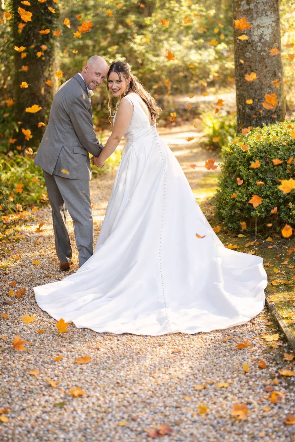 Bride in white gown and groom in gray suit walk hand-in-hand on a gravel path surrounded by fall foliage.
