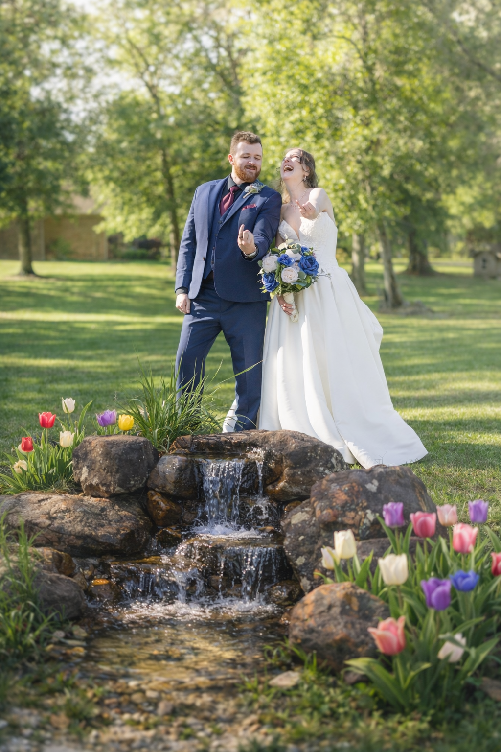 Joyful bride and groom posing by a stone waterfall surrounded by colorful tulips in a lush green park.