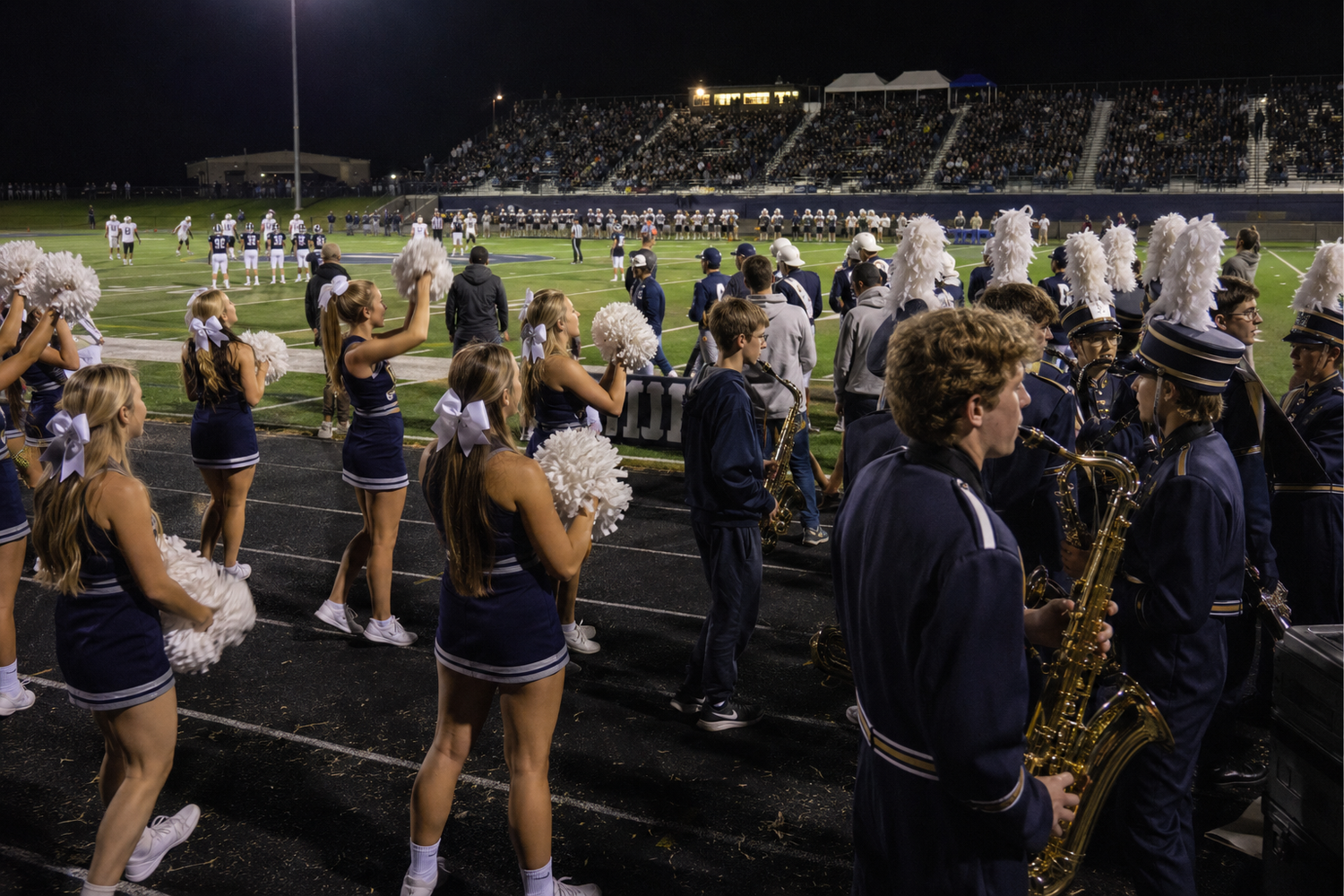 Cheerleaders with pom-poms and marching band members in navy uniforms perform on sidelines during a night football game.