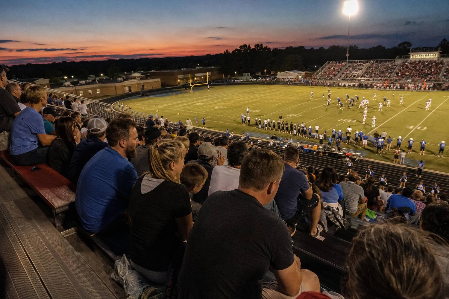 Fans watch a high school football game at dusk from packed bleachers with a colorful sunset sky backdrop.