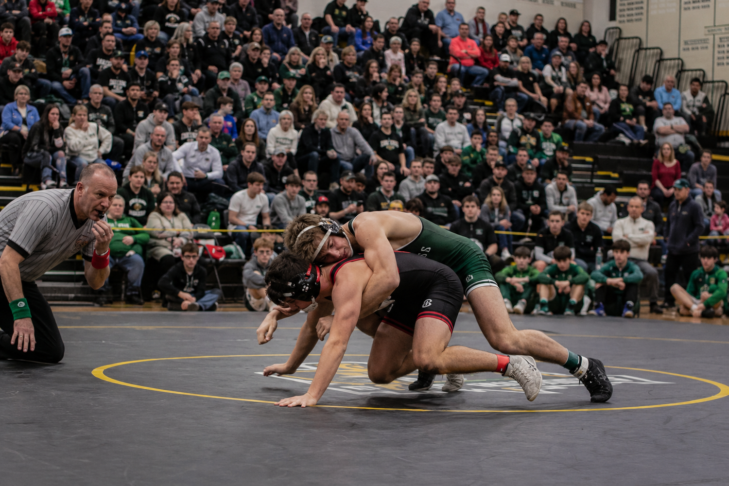 Two wrestlers compete on mat as referee watches closely during an indoor wrestling match with packed gymnasium crowd.