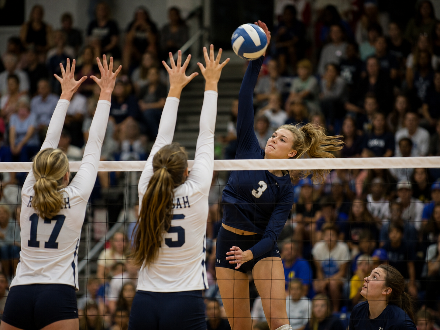 Volleyball player number 3 spikes the ball over two blockers during a competitive high school match.