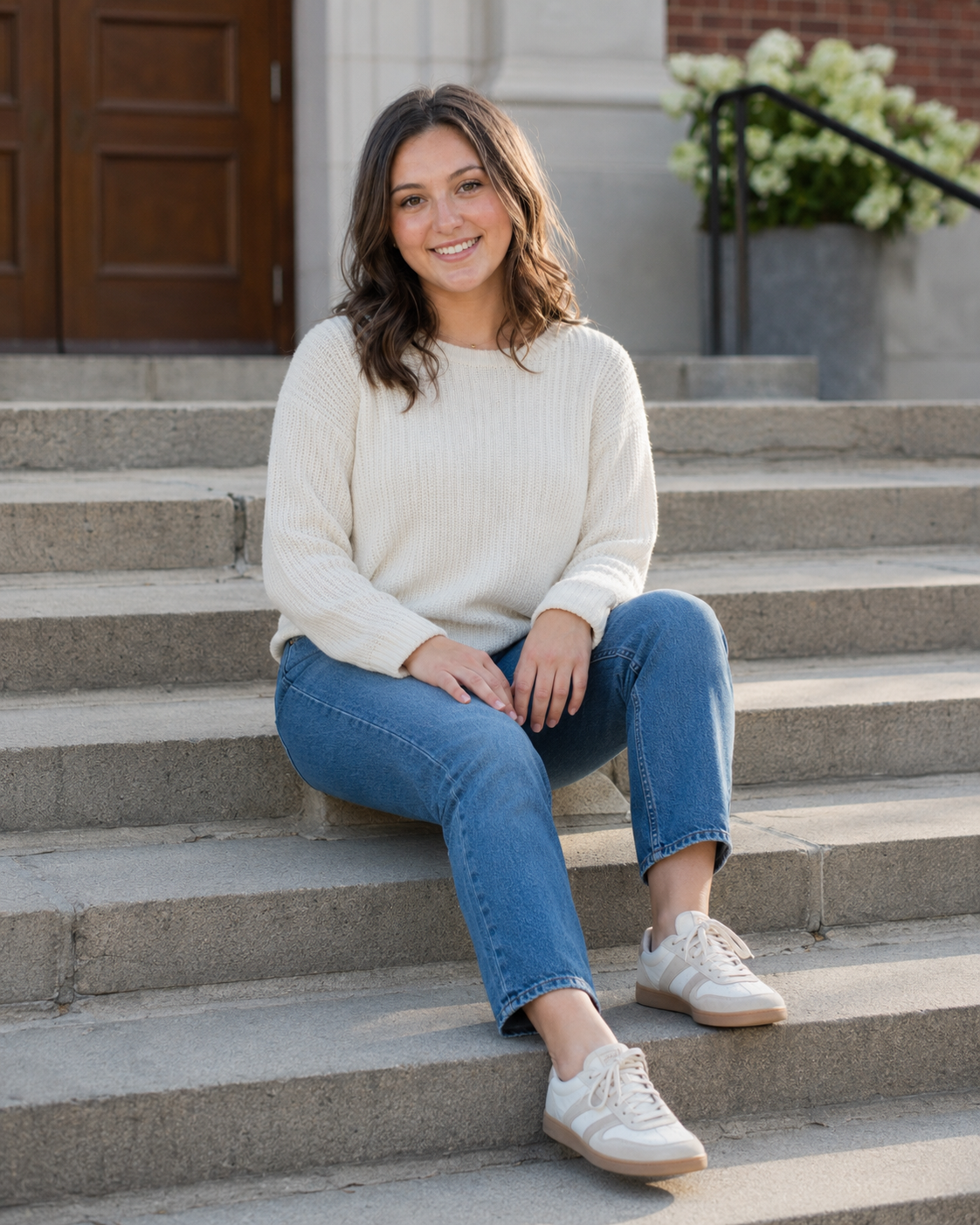 Smiling young woman in cream sweater and jeans sitting on stone steps outdoors.
