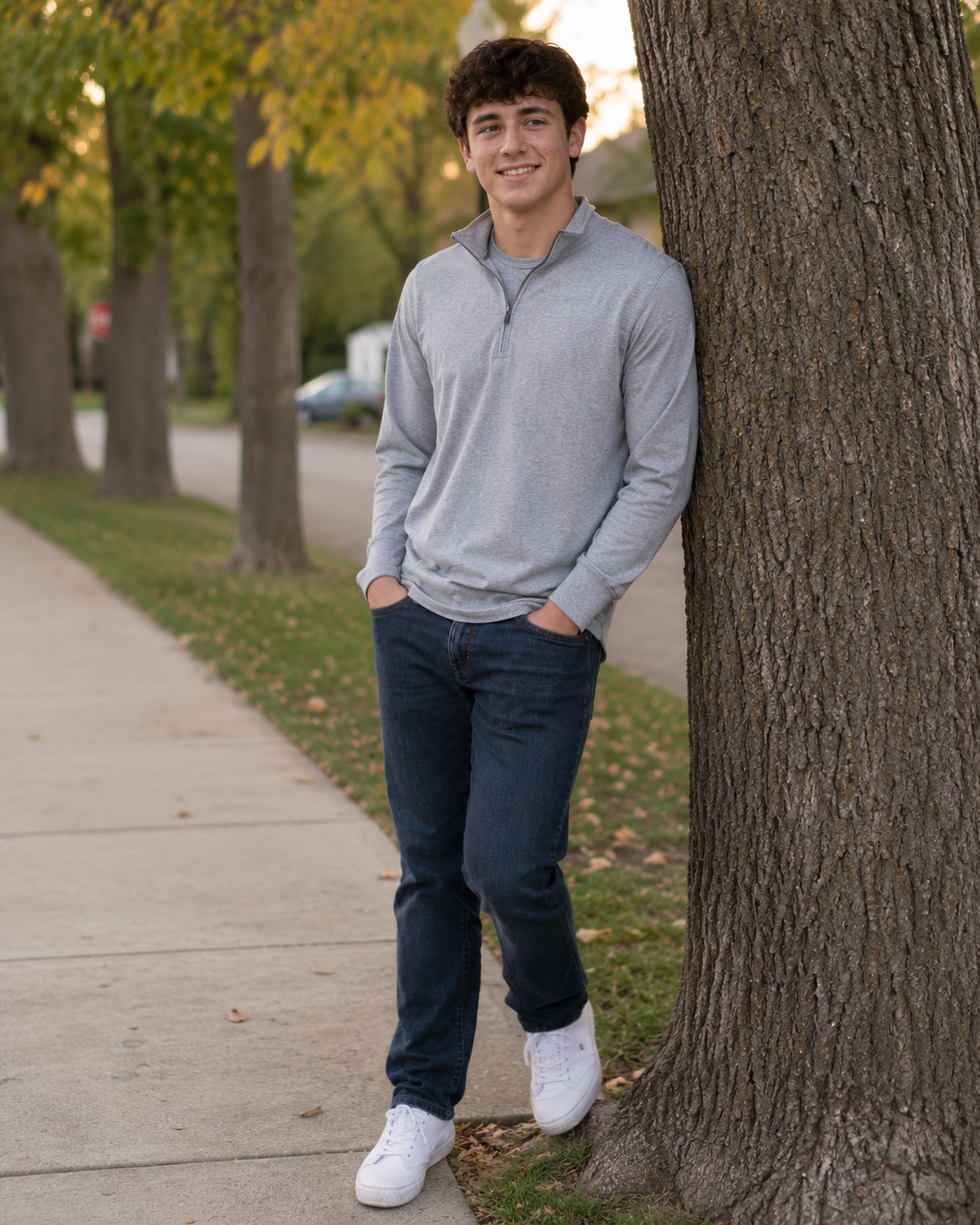 Young man in gray quarter-zip leaning against a tree on a sidewalk in autumn setting.