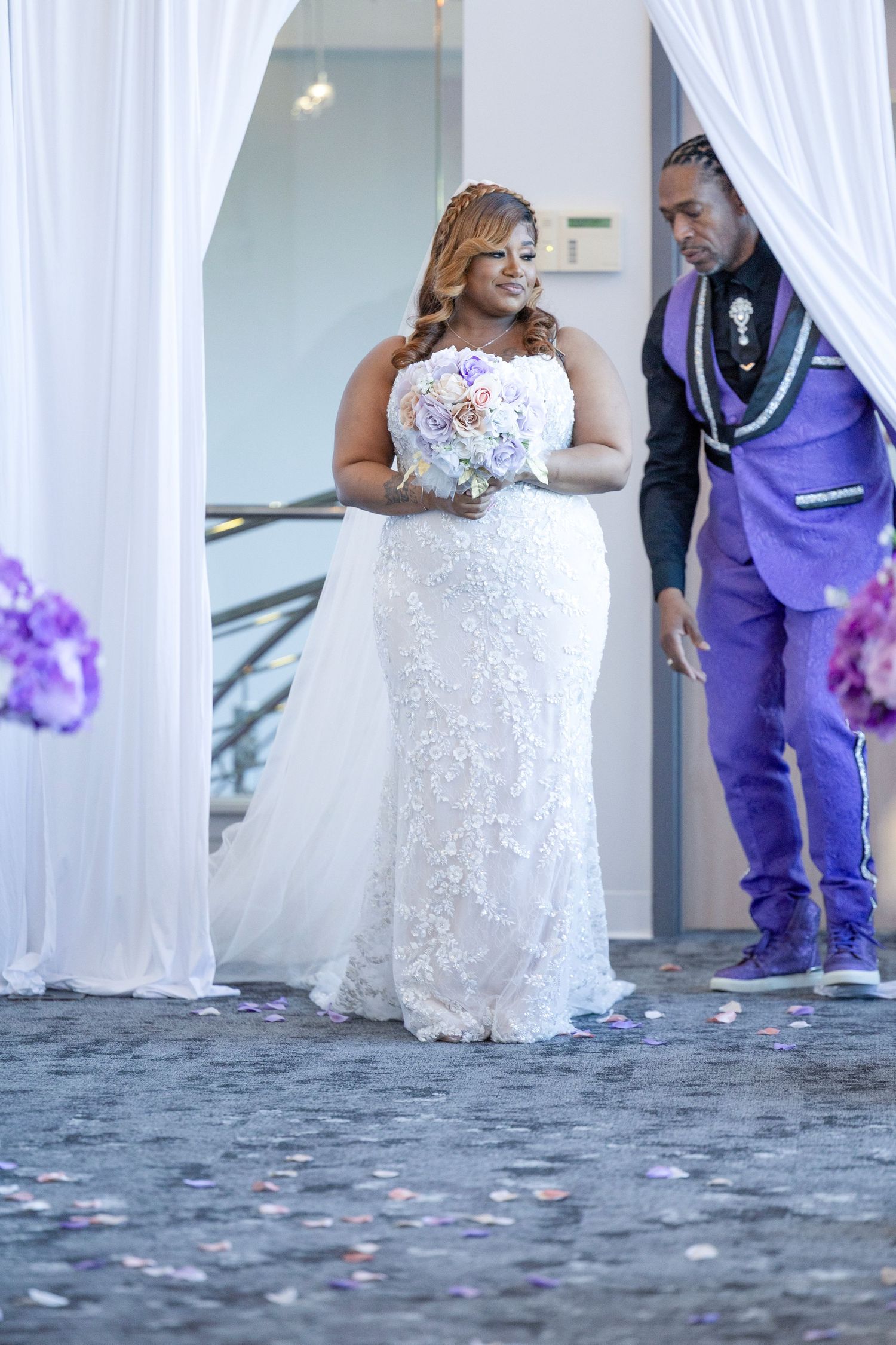 Bride in white lace gown holds pastel bouquet beside groom in purple suit at elegant wedding ceremony.