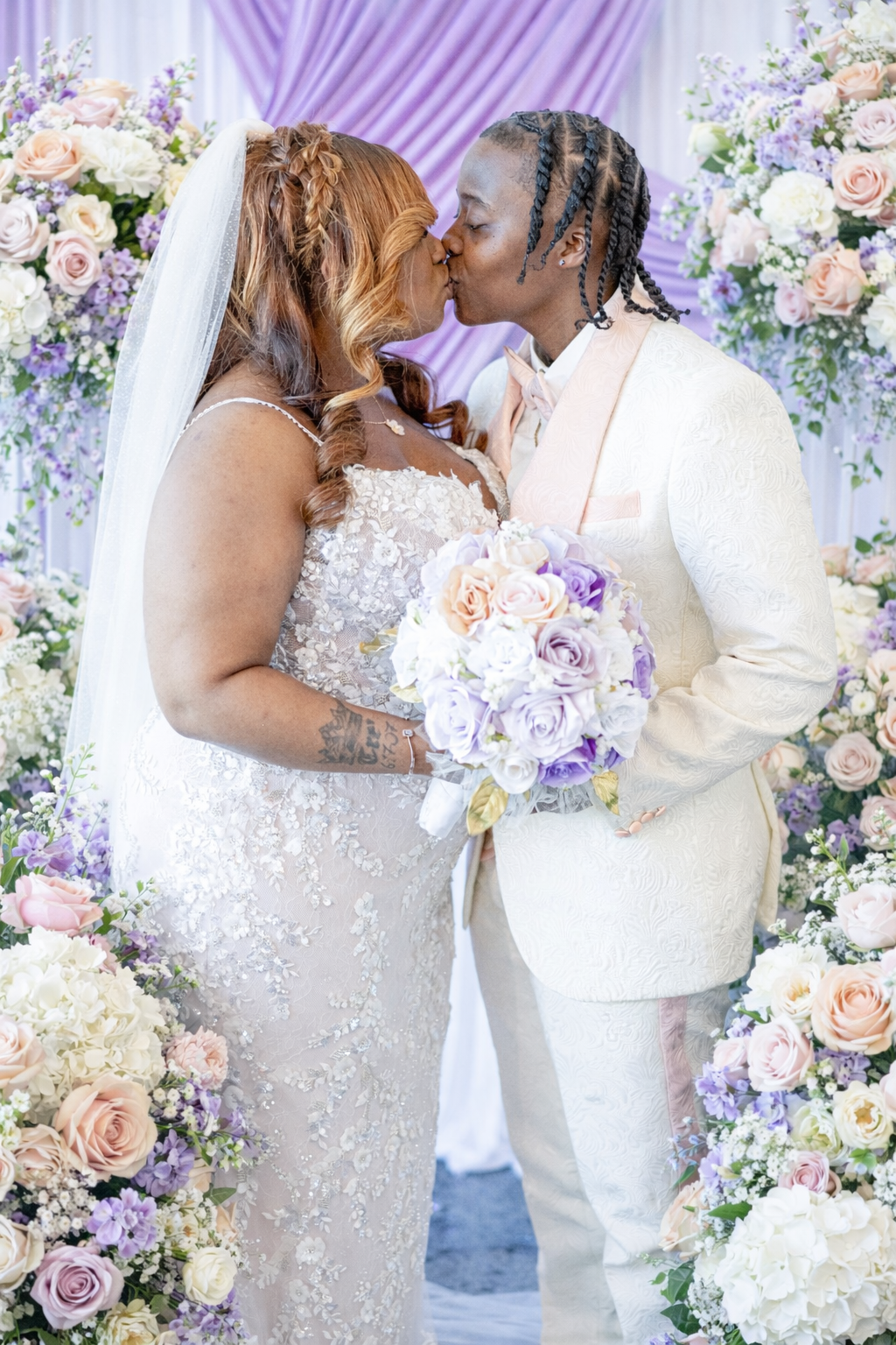 Two brides share a kiss at their wedding altar surrounded by lavender and blush floral arrangements.