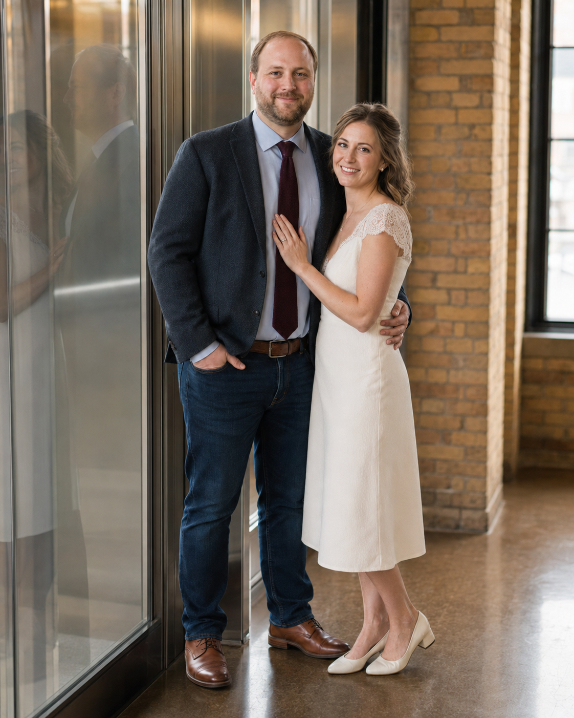 A smiling couple poses together, man in navy blazer and burgundy tie, woman in white midi dress, in a brick-walled venue.