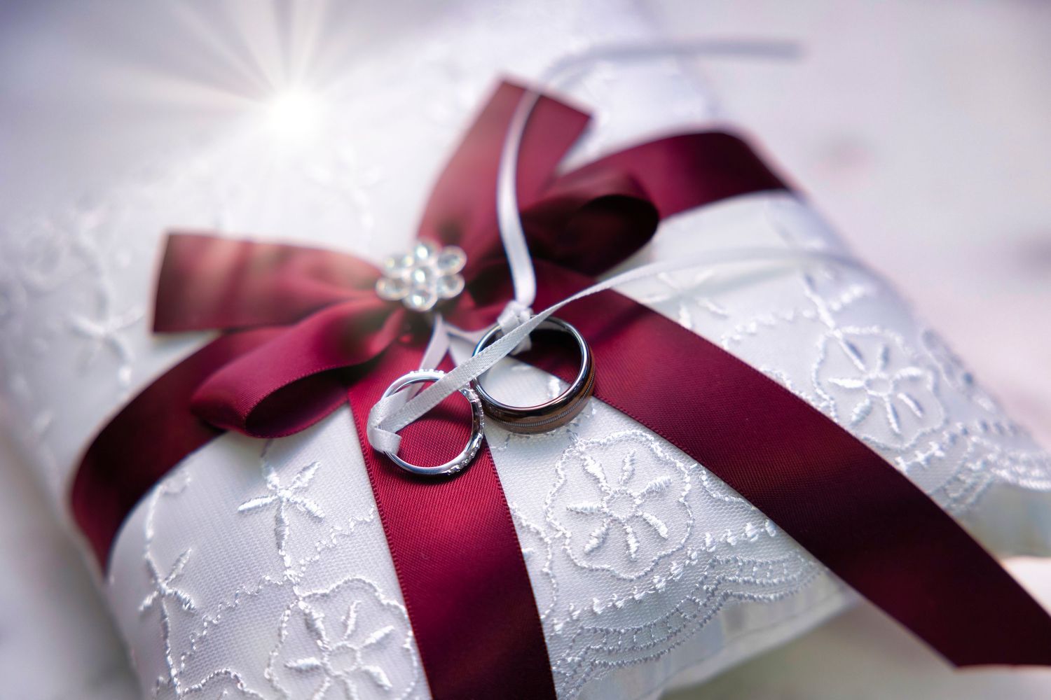 Wedding rings rest on a white lace pillow with burgundy ribbon decoration.