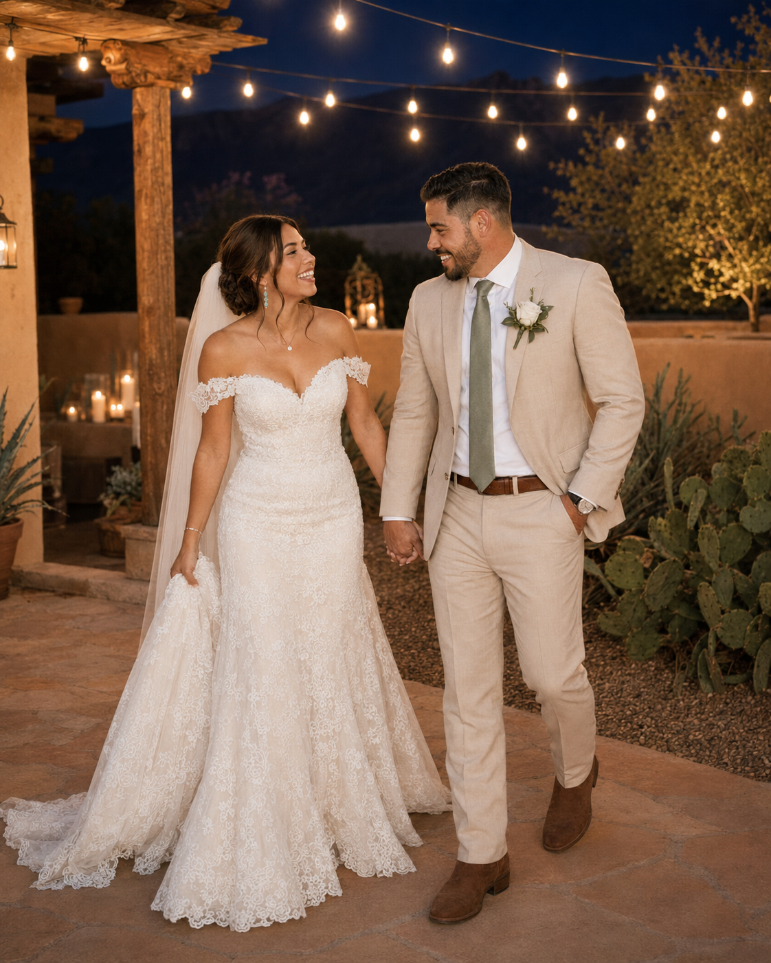 A bride and groom hold hands smiling at each other outdoors at night with string lights glowing behind them.