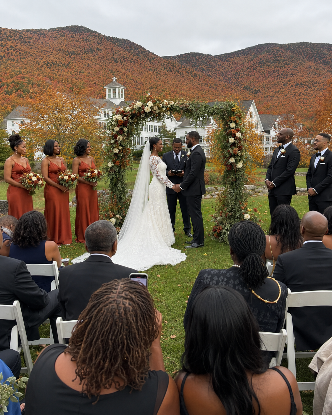 Outdoor fall wedding ceremony with floral arch, bride and groom, bridesmaids in rust dresses, mountain foliage backdrop.