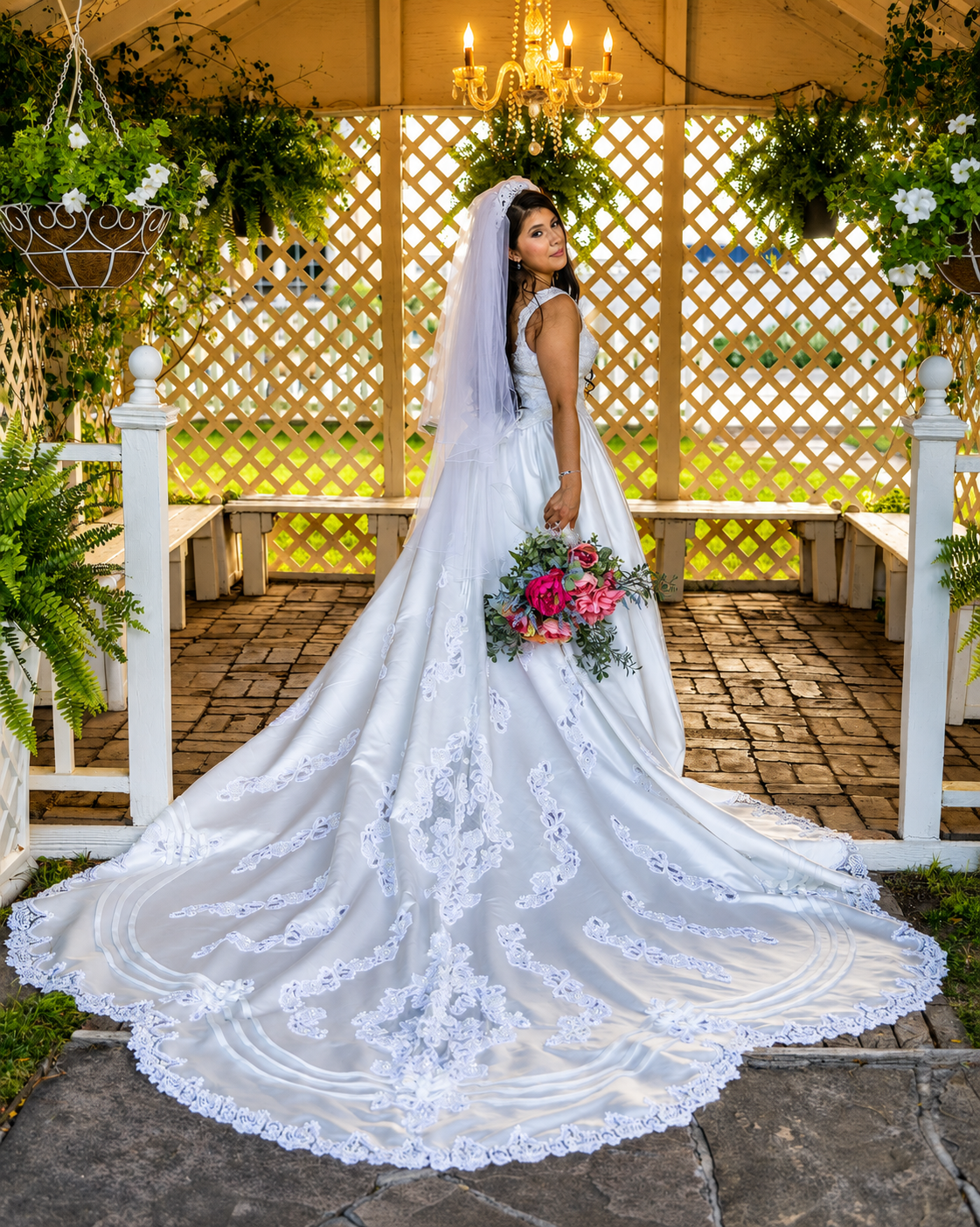 Bride in elegant lace wedding gown with long cathedral veil holds pink bouquet in a white gazebo.