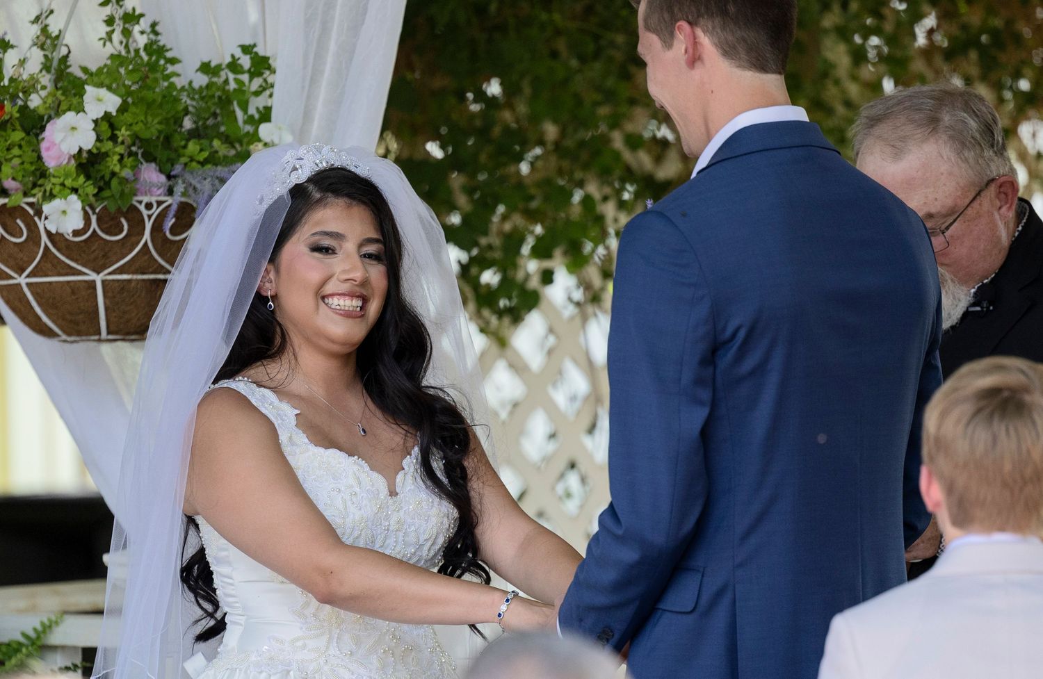 Smiling bride in white gown and veil holds hands with groom in blue suit during outdoor wedding ceremony.