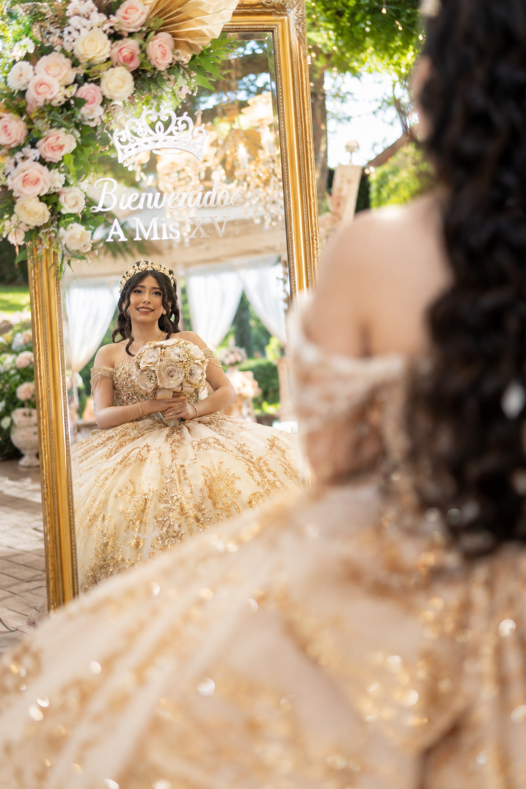 Quinceañera girl in gold ballgown reflected in floral-adorned mirror reading Bienvenidos A Mis XV.