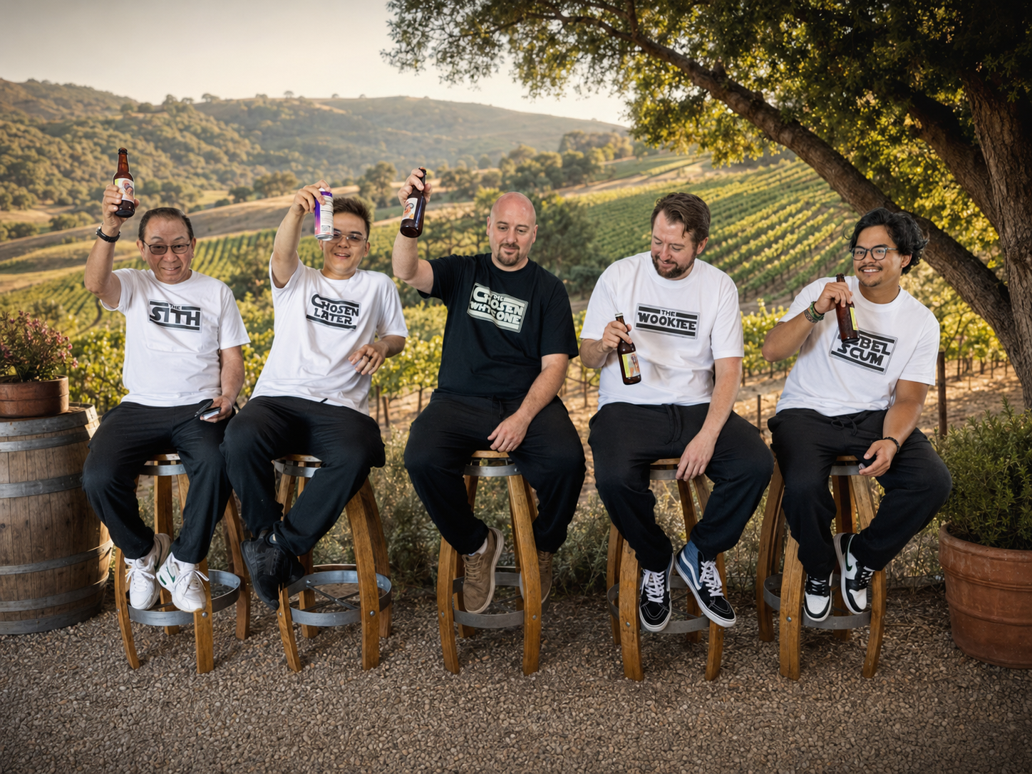 Five friends in white t-shirts celebrate with drinks on stools amid scenic vineyard hills at golden hour.