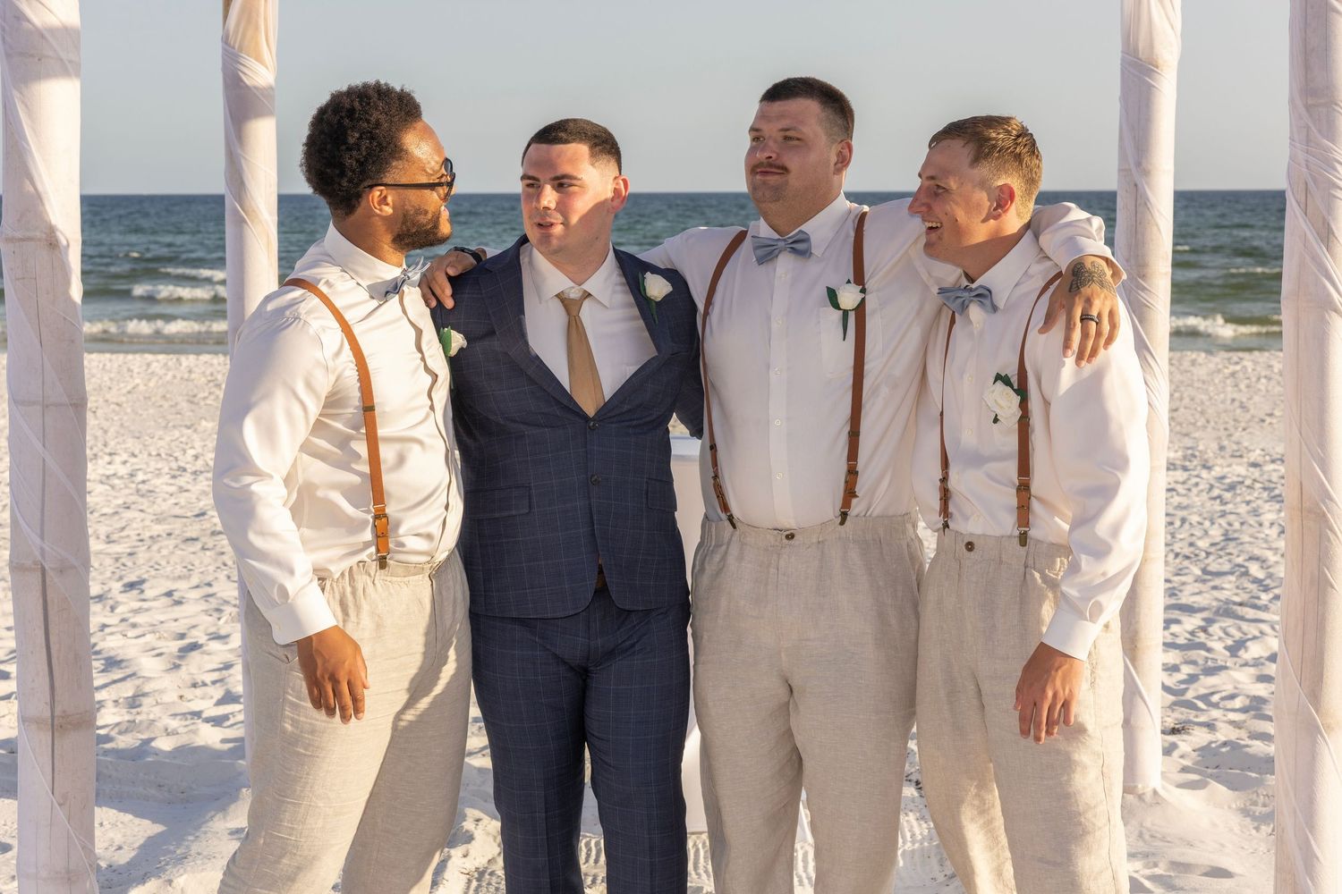 Groom in navy suit with three groomsmen in tan suspenders posing on a beach wedding ceremony.