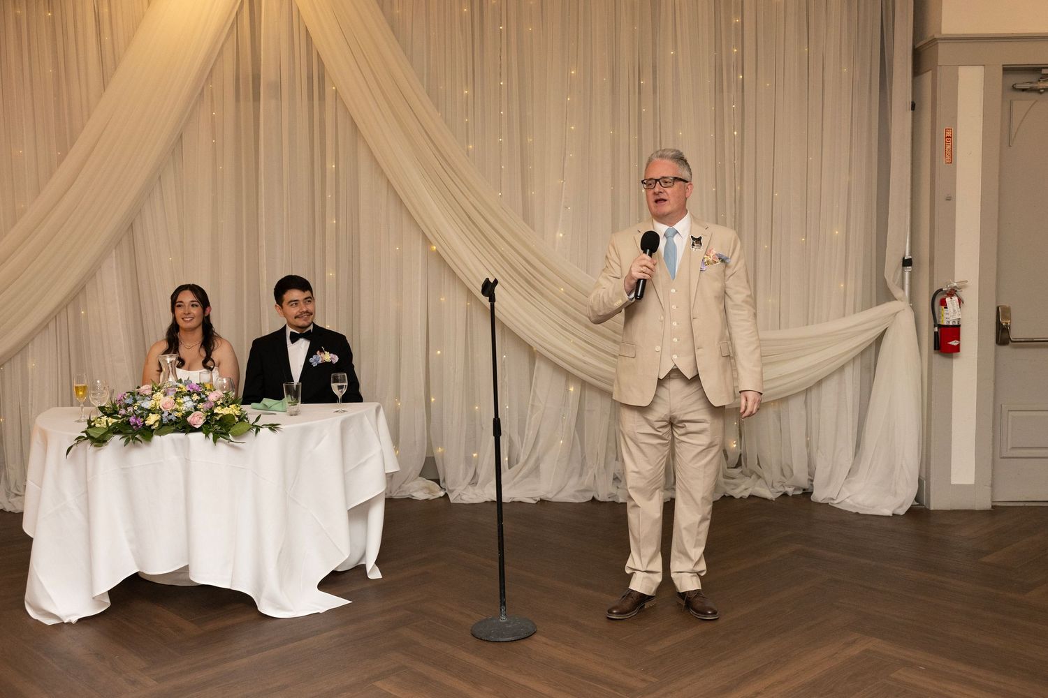 Man in beige suit giving wedding speech at microphone while bride and groom sit at sweetheart table.