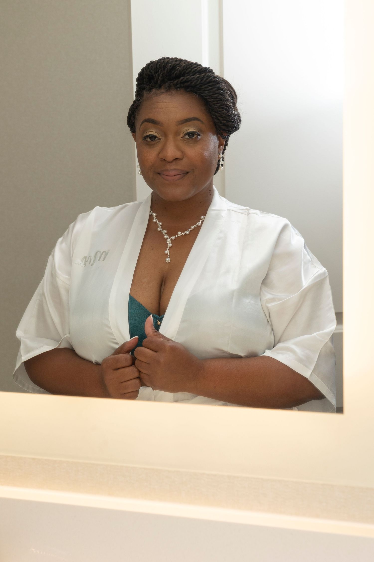 Woman in white robe and teal bra smiling at mirror, wearing diamond necklace and earrings.