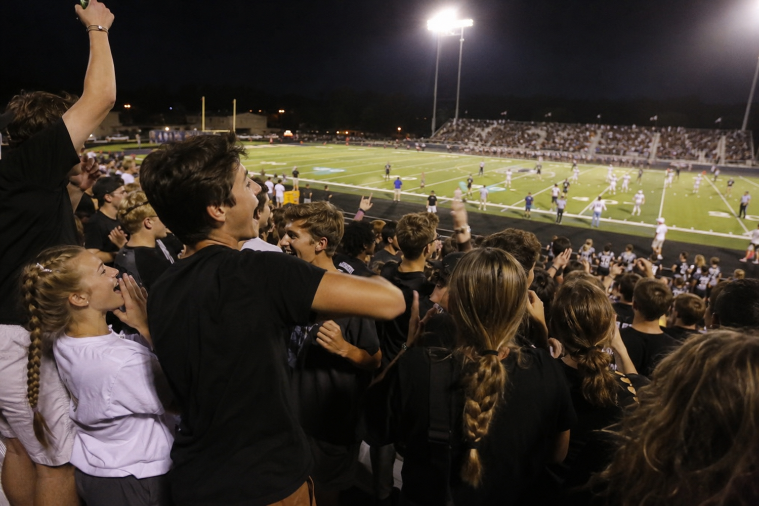 Excited crowd of students in black shirts cheering at a night high school football game under bright stadium lights.