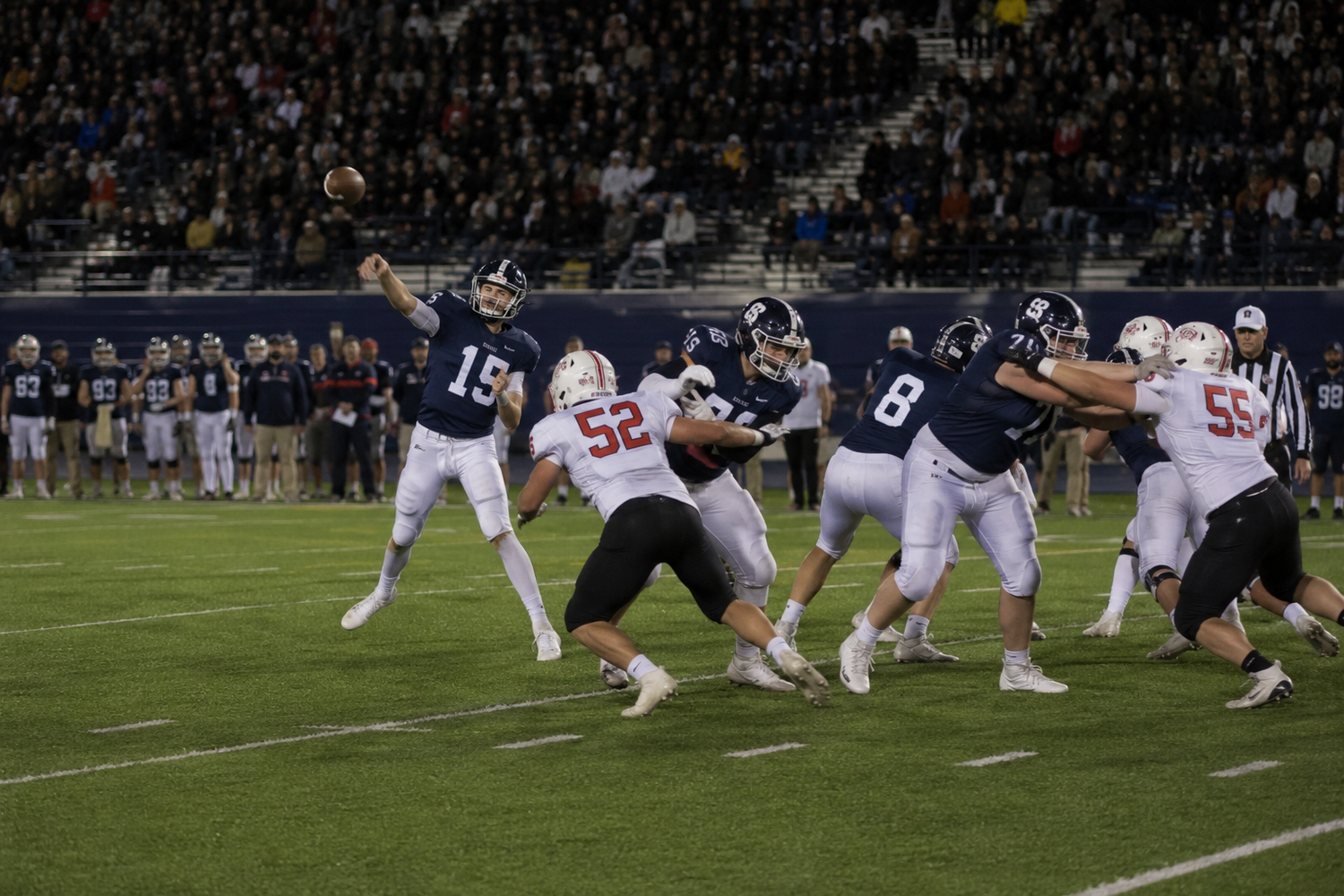 College football quarterback #19 throws a pass while offensive linemen block defenders during a night game.