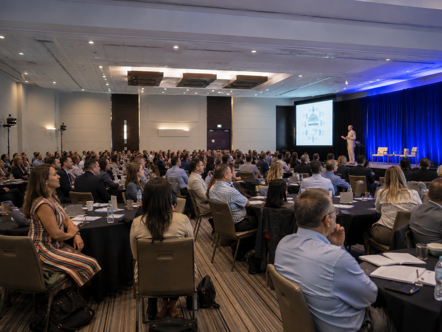 A large audience seated at round tables listens to a speaker presenting on stage at a professional conference.