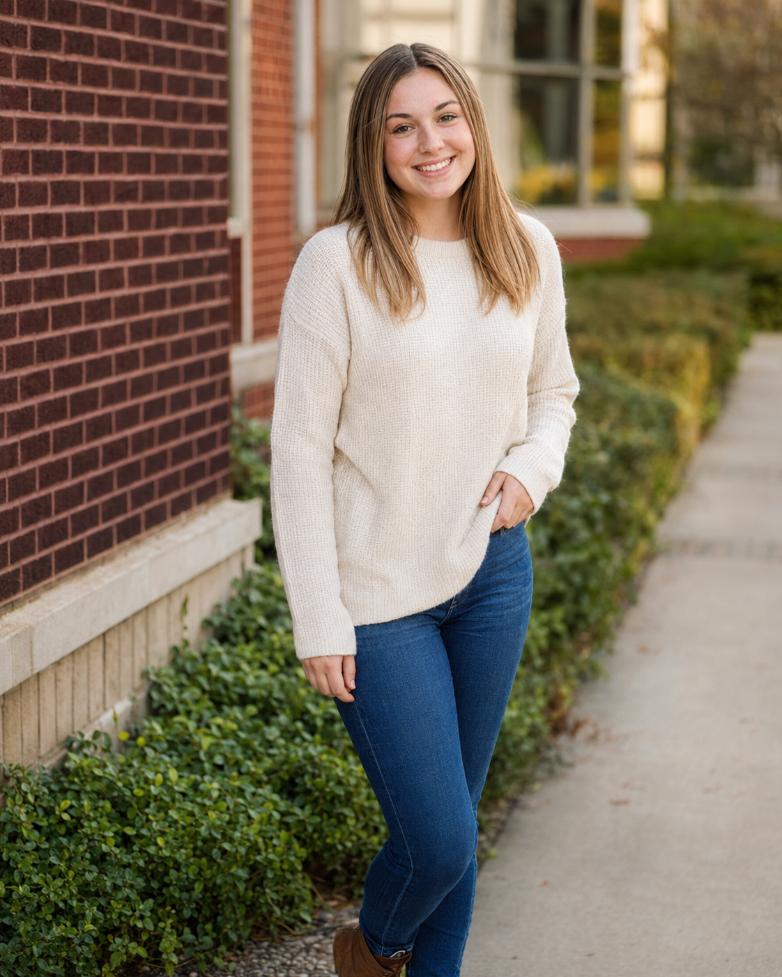 Smiling young woman in cream sweater and jeans posing outdoors near a brick building with green hedges.