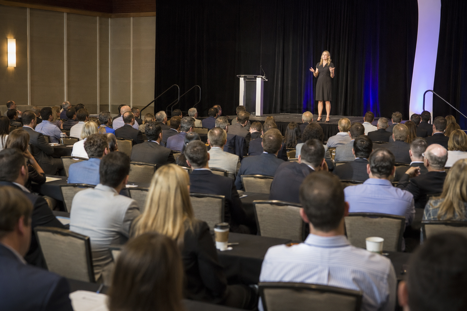 A blonde speaker presents on stage to a large audience at a professional conference event.