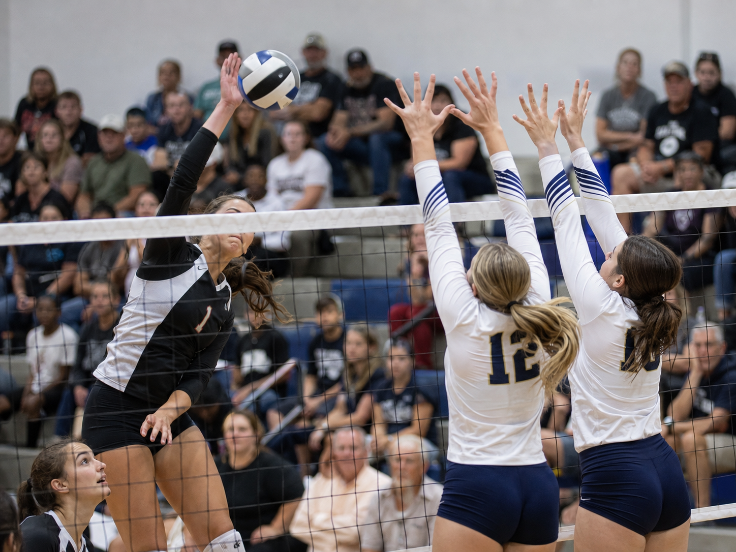 Volleyball players compete at the net during an intense high school match with fans watching.