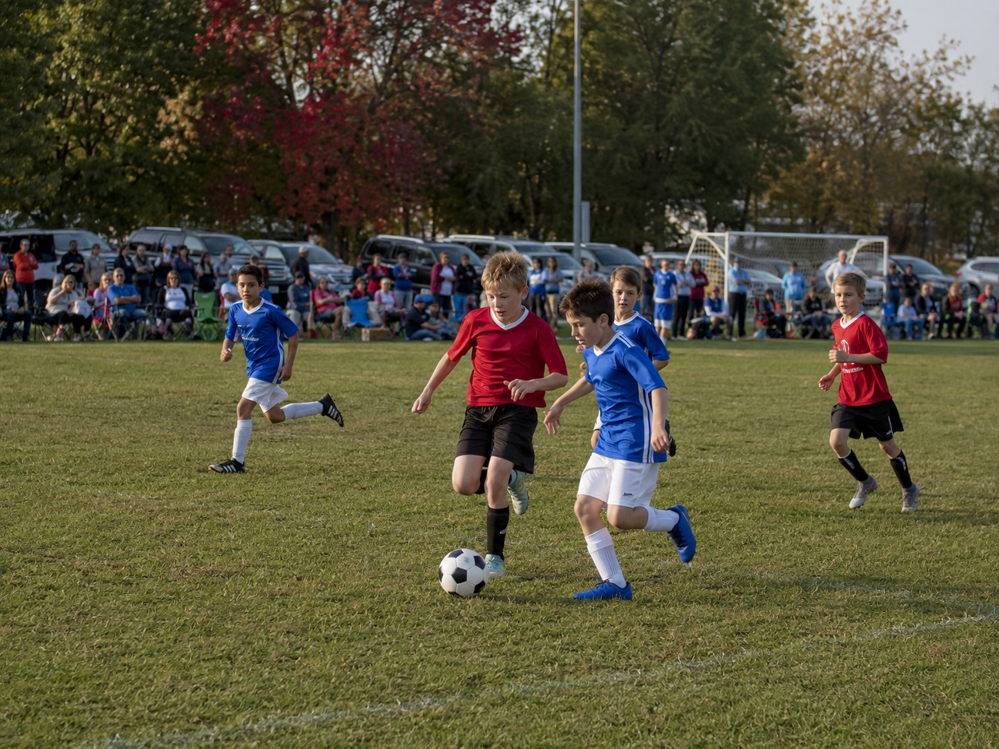 Youth soccer players in red and blue uniforms compete for the ball on a green field in autumn.