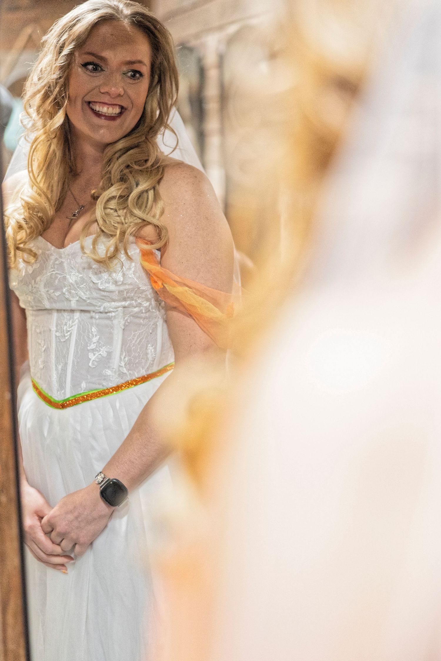 Smiling blonde bride in white strapless gown with gold belt and orange sash, wearing a smartwatch, posing elegantly.