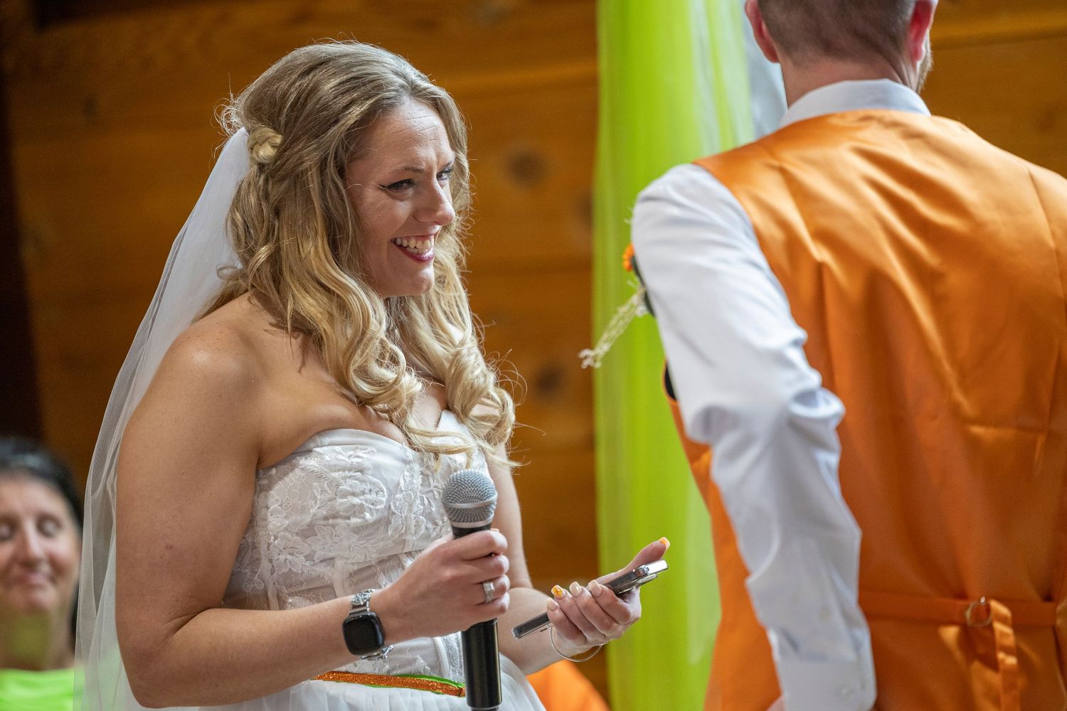 Smiling bride in white lace gown holding microphone during wedding ceremony with colorful backdrop.