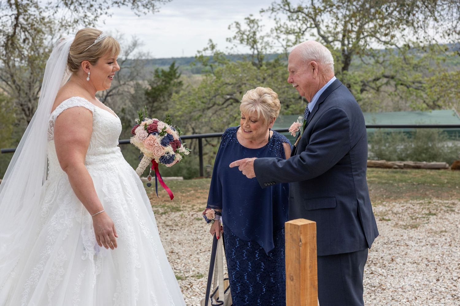 Bride in white gown holds bouquet while elderly couple adjusts boutonniere outdoors at rustic wedding venue.