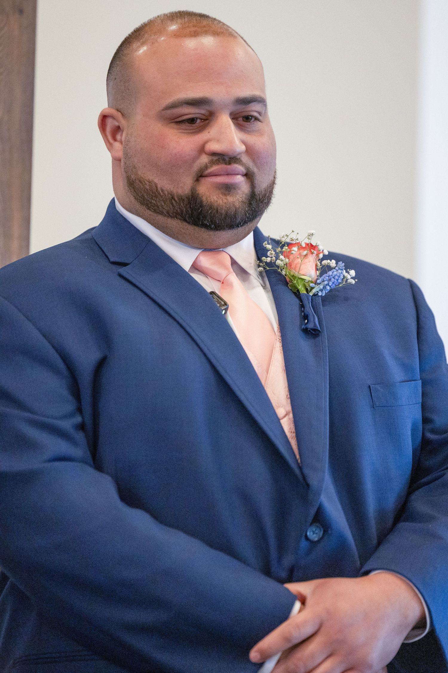Man in navy blue suit with pink tie and floral boutonniere smiling at a wedding ceremony.