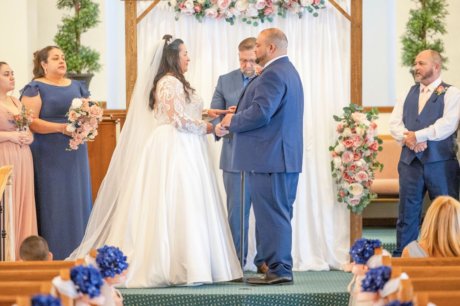 Bride and groom exchanging rings at church altar with bridesmaids in blue dresses and officiant during wedding ceremony.