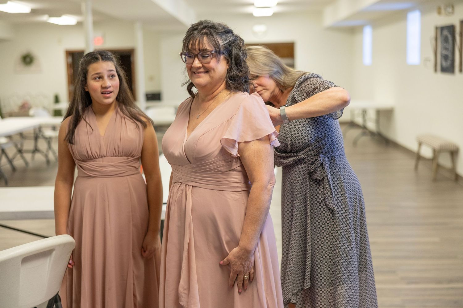 Three women in mauve bridesmaid dresses stand in a hall, with one woman being embraced from behind.