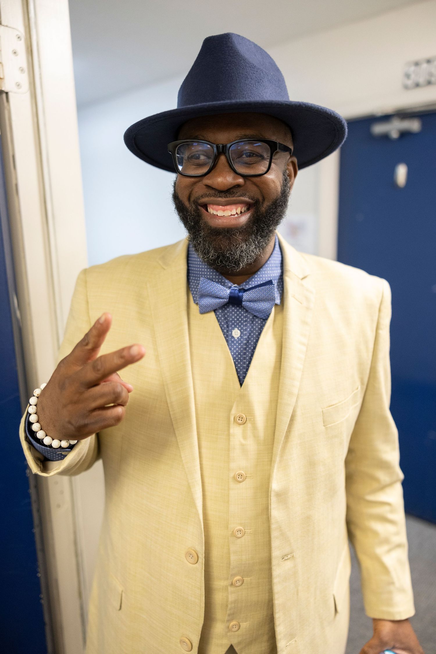 Smiling man in yellow suit, blue bow tie, navy fedora hat, and glasses making a hand gesture.