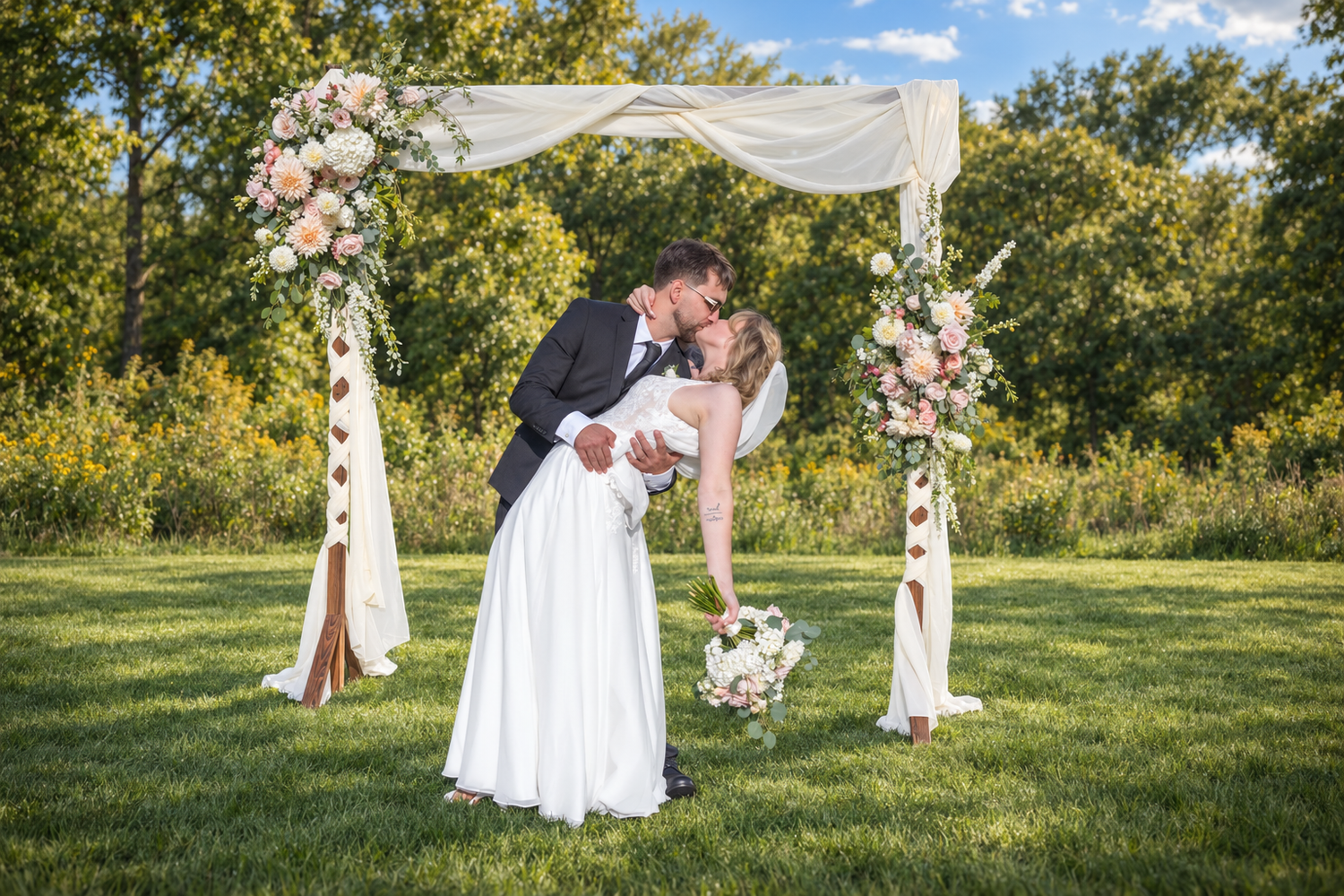 Bride and groom sharing a romantic kiss under a floral arch adorned with white draping and pink roses outdoors.