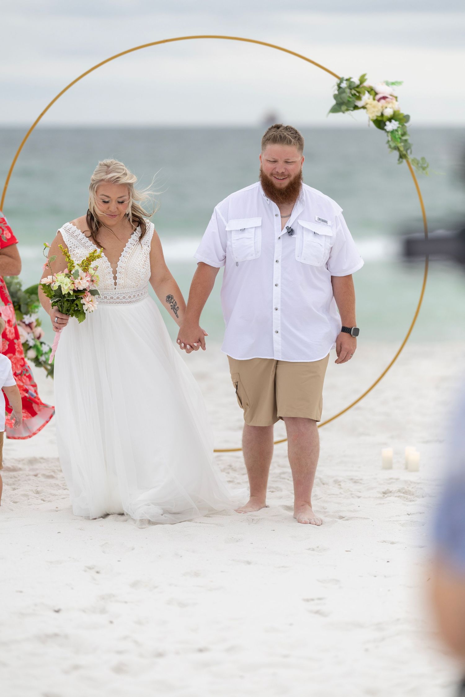 Bride and groom hold hands at beach wedding ceremony under gold circular arch with flowers, ocean backdrop.