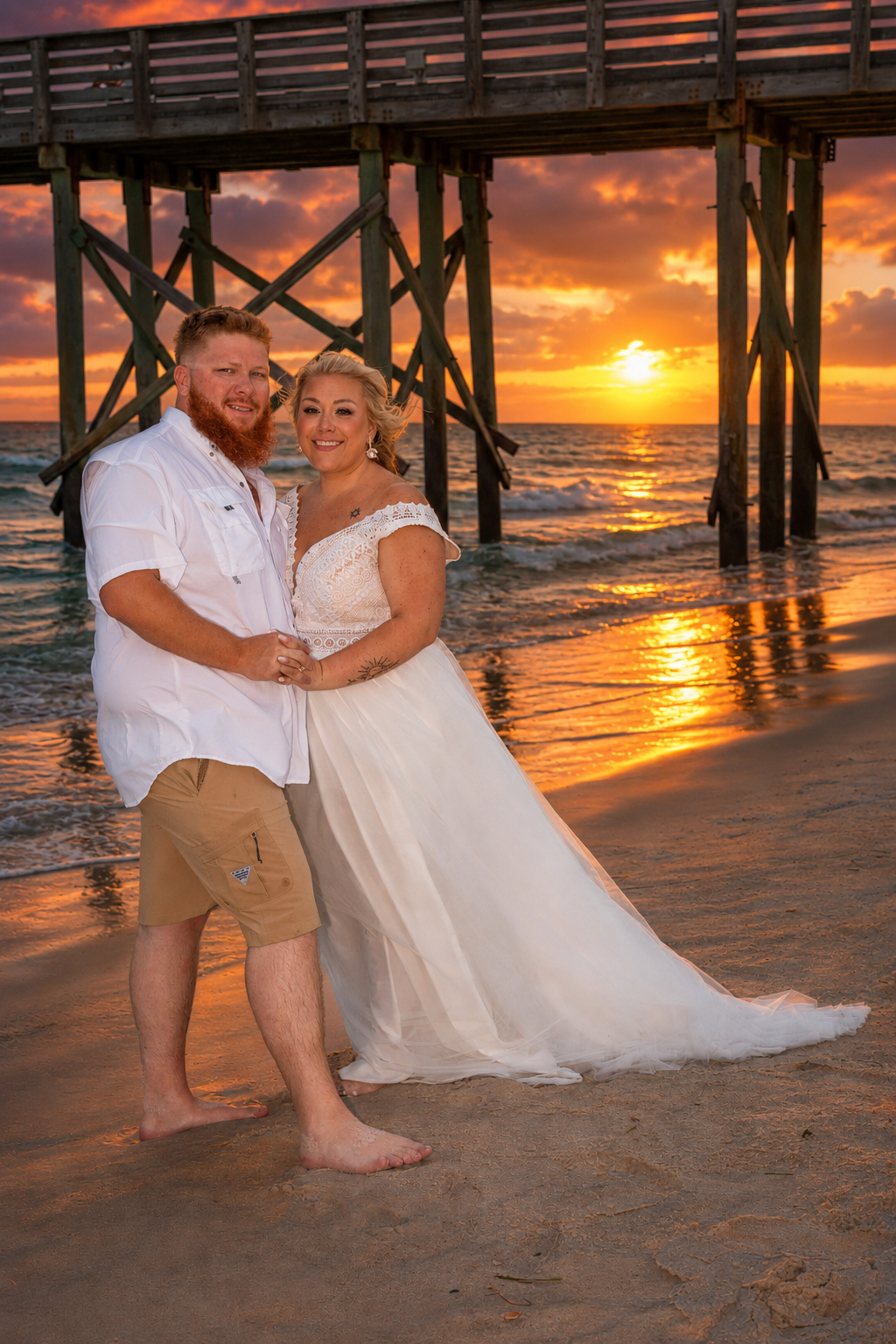 Bride and groom embracing barefoot on a beach at golden sunset with a wooden pier in the background.