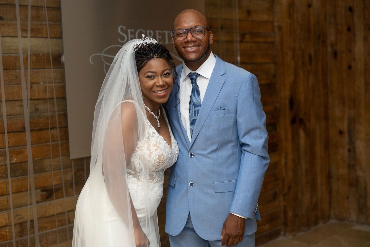 A happy couple poses together at their wedding, with the bride in a white lace gown and the groom in a light blue suit.